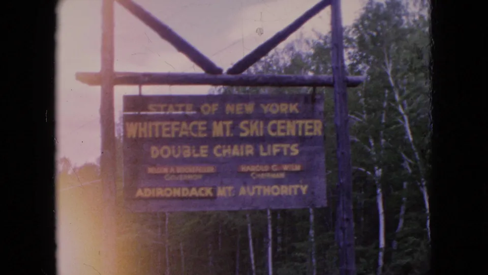 A 1964 8mm frame shows a painted State of New York Whiteface Mountain Ski Center sign for double chair lifts, mounted on rustic posts with trees behind it.
