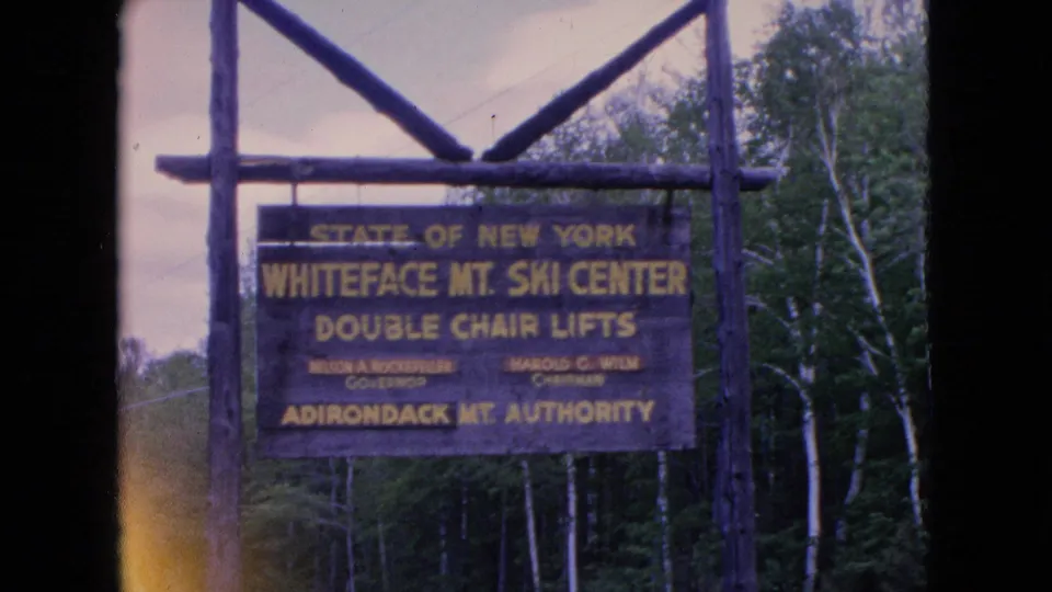 1964 8mm frame showing a wooden Whiteface Mountain Ski Center sign for double chair lifts mounted on rustic posts in a wooded New York mountain setting.