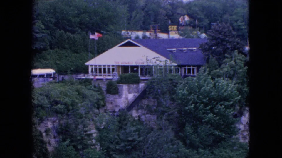 Wide 8mm view of the Whiteface Mountain lodge area, with a sloped-roof building, large windows, an American flag, trees, and rocky cliffside terrain.