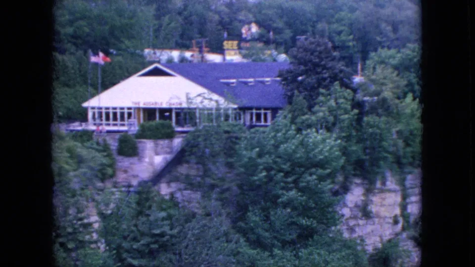 1964 home-movie frame of the Whiteface Mountain lodge seen across dense greenery, with the building set above a rock face and partially obscured by trees.
