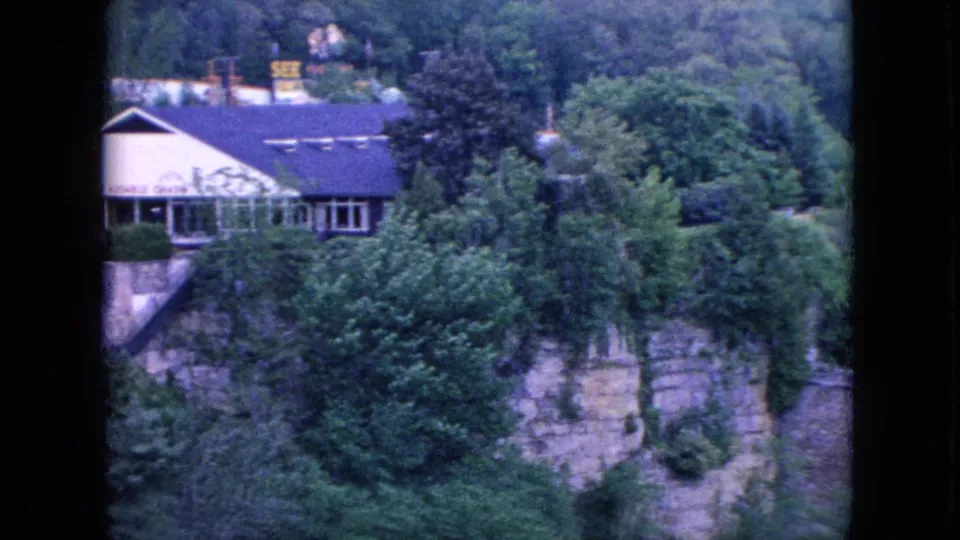 Soft 8mm frame showing part of the Whiteface Mountain lodge and surrounding Adirondack greenery above a rocky slope, with roadside signage faintly visible in the background.