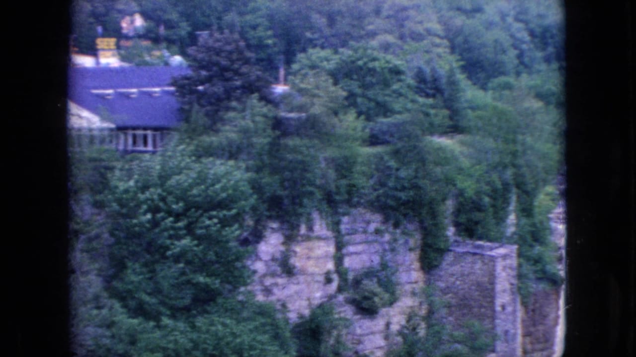 1964 8mm home-movie frame from the Niagara Falls, New York area showing dense woods, stone or masonry walls, and a house-like structure above the river gorge.