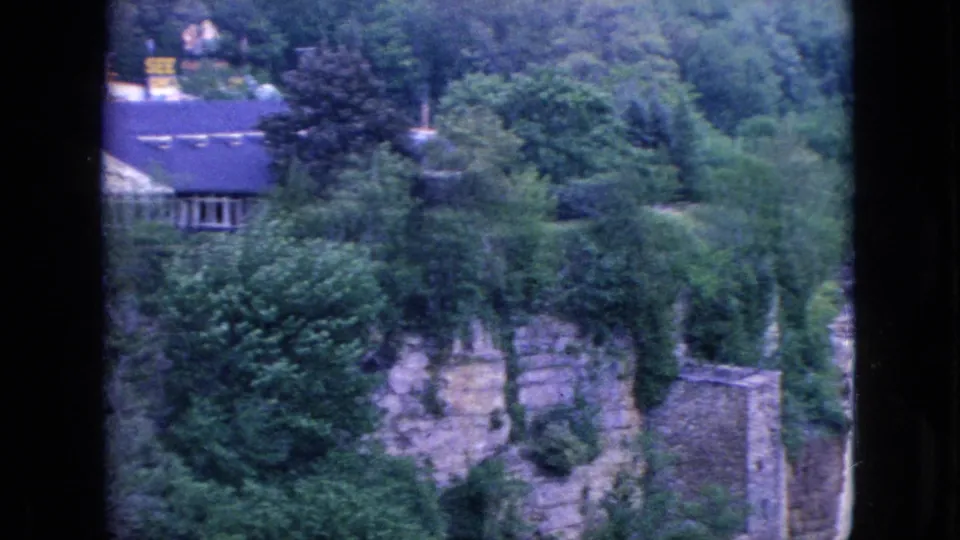 1964 8mm home-movie frame from the Niagara Falls, New York area showing dense woods, stone or masonry walls, and a house-like structure above the river gorge.