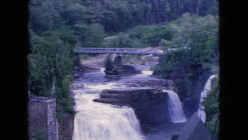 1964 8mm frame of a waterfall dropping through a rocky Niagara Falls-area gorge, with a bridge above and dense greenery along the banks.