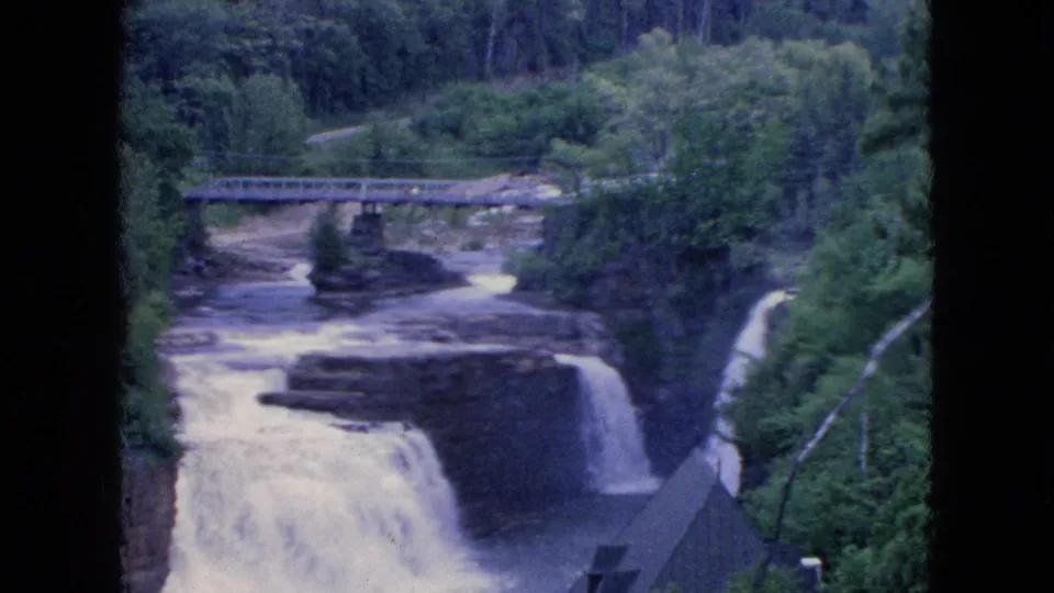 1964 Niagara Falls-area home-movie frame showing a wide scenic view of a bridge over rocky whitewater and a waterfall framed by dense green foliage.
