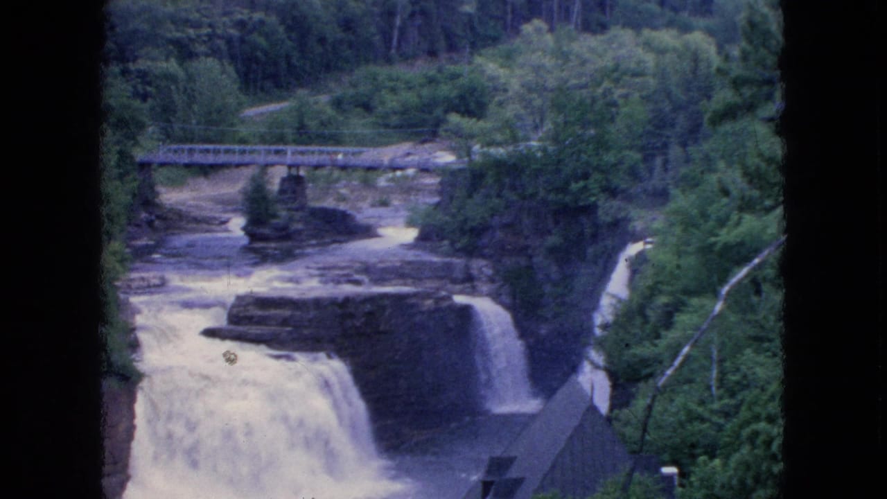 1964 8mm visitor footage from the Niagara Falls, New York area shows a river passing beneath a metal bridge and dropping over rocky ledges amid dense trees.