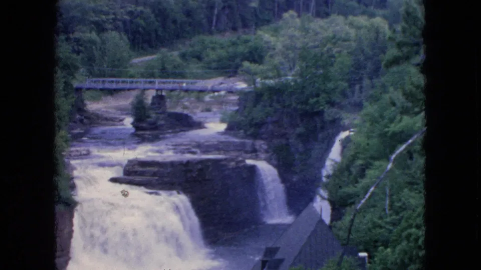 1964 8mm visitor footage from the Niagara Falls, New York area shows a river passing beneath a metal bridge and dropping over rocky ledges amid dense trees.