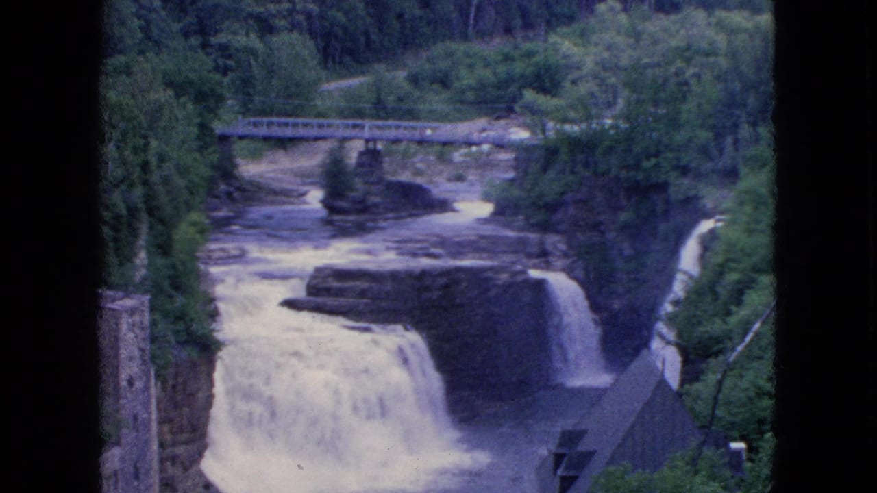 Water flows over multiple falls below a bridge with wooded banks and a roof at lower right.