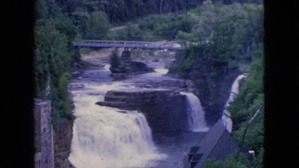 The Niagara Falls, New York area cascade appears in soft 8mm color, with white water dropping over rock shelves below a bridge and wooded banks.