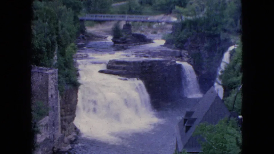 A wider 1964 8mm frame shows the waterfall descending toward the lower river, with a stone structure at left, a bridge above, trees, and dark film edges.