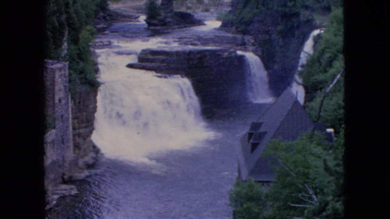 1964 8mm Niagara Falls travel frame showing white water dropping over rocky ledges beside trees, a stone tower, and a steep roofed riverside structure.