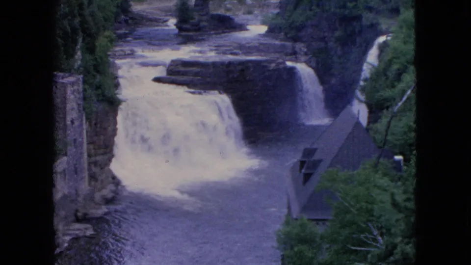 1964 8mm Niagara Falls travel frame showing white water dropping over rocky ledges beside trees, a stone tower, and a steep roofed riverside structure.
