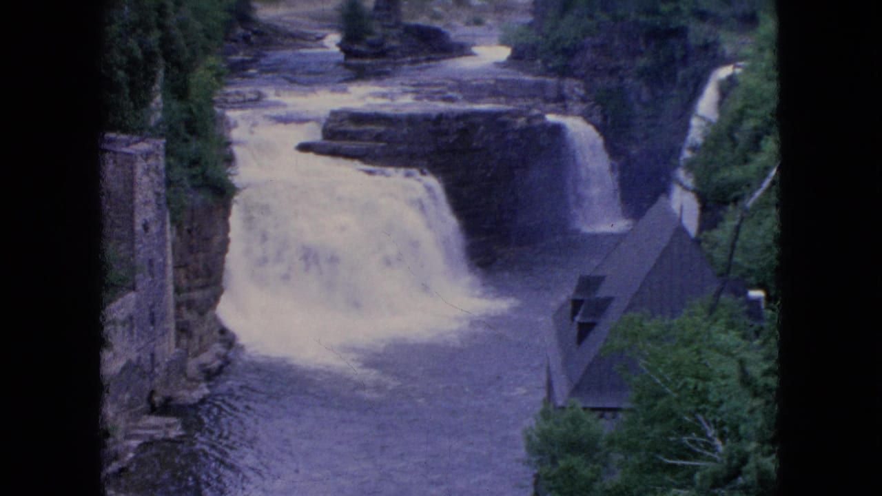 1964 8mm Niagara Falls frame with white rushing water, layered rock, surrounding trees, a stone tower, and a steep roofed structure near the river.