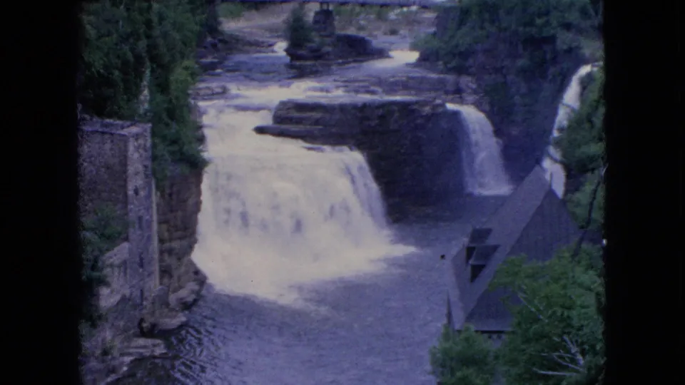 1964 8mm Niagara Falls home-movie frame showing a broad waterfall, dark river current, wooded banks, a stone tower, and nearby roofed structure.