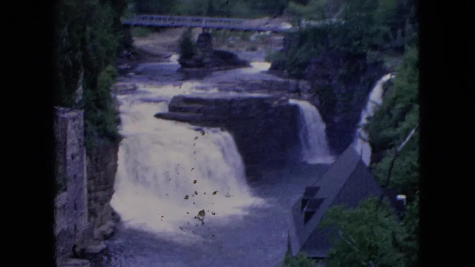1964 8mm Niagara Falls frame showing cascading water, a bridge above the river, wooded banks, a stone tower, and a steep roofed structure beside the falls.