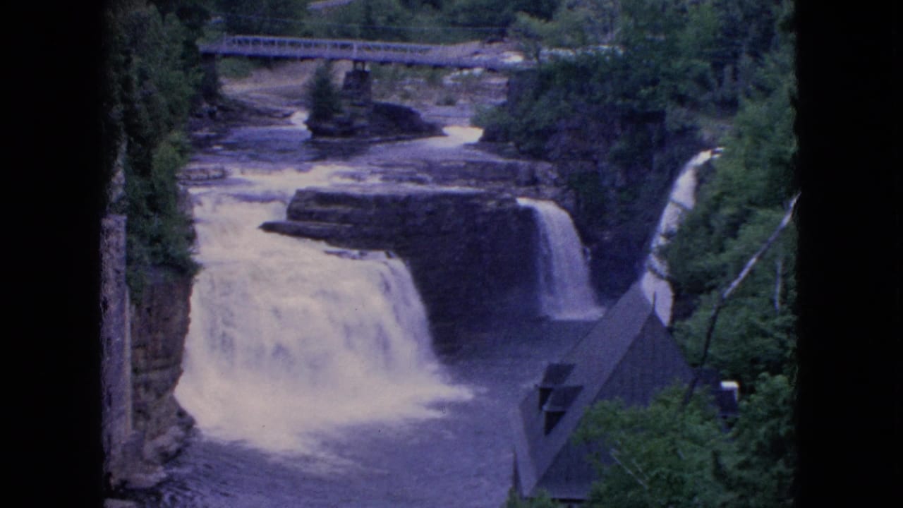 1964 8mm home-movie frame from Niagara Falls, New York metadata showing a waterfall dropping through a rocky, tree-lined gorge with a bridge in the background.