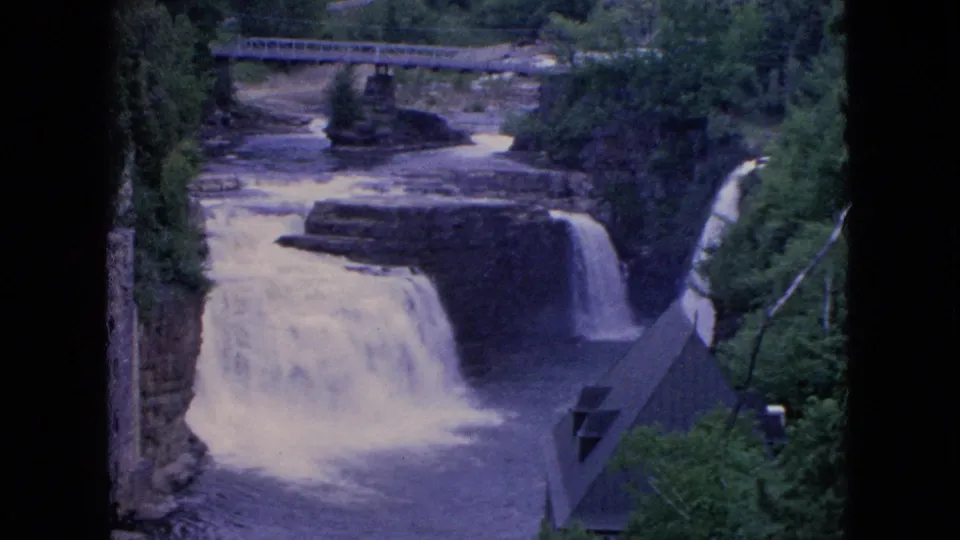 1964 8mm home-movie frame from Niagara Falls, New York metadata showing a waterfall dropping through a rocky, tree-lined gorge with a bridge in the background.