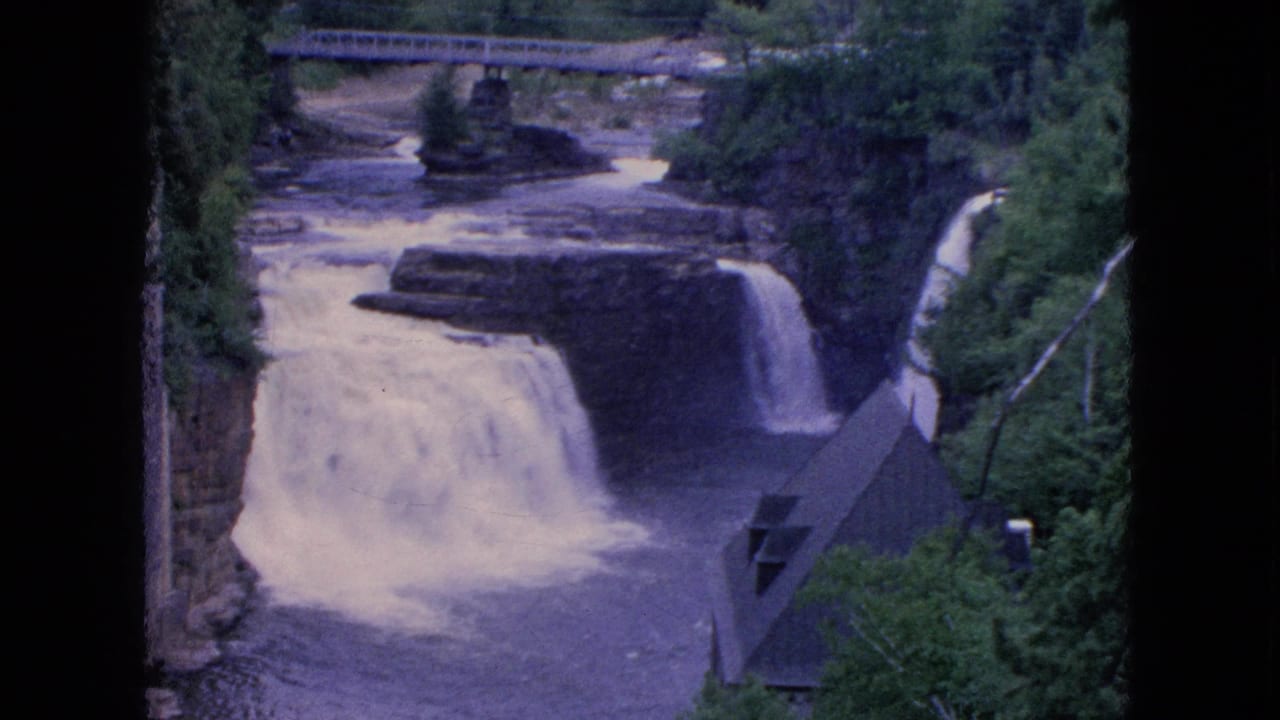 Rocky waterfall scene with dense trees, bridge above, and dark-roofed structure near the water.