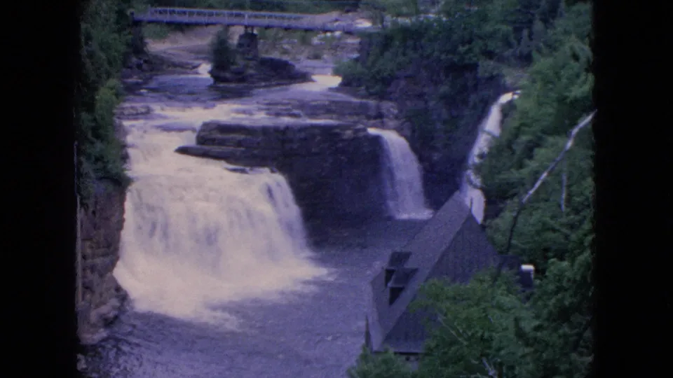 1964 8mm home-movie frame from Niagara Falls, New York metadata showing a steady scenic view of a waterfall, surrounding rock walls, thick trees, and a bridge upriver.
