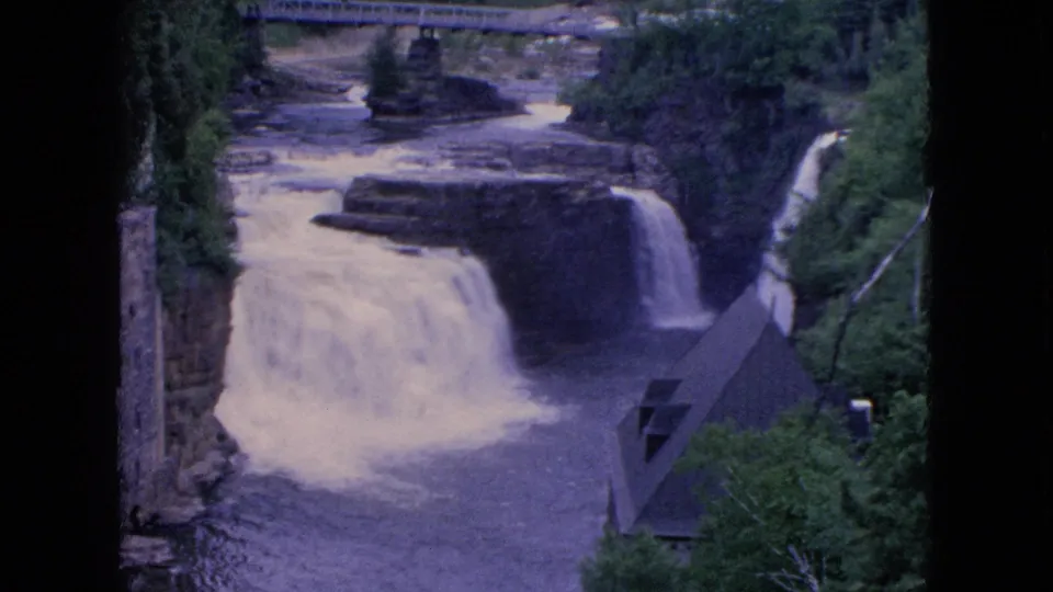 1964 8mm home-movie frame from Niagara Falls, New York metadata showing the waterfall at the end of the clip, with wooded banks, rocky ledges, and a bridge crossing the upper waterway.