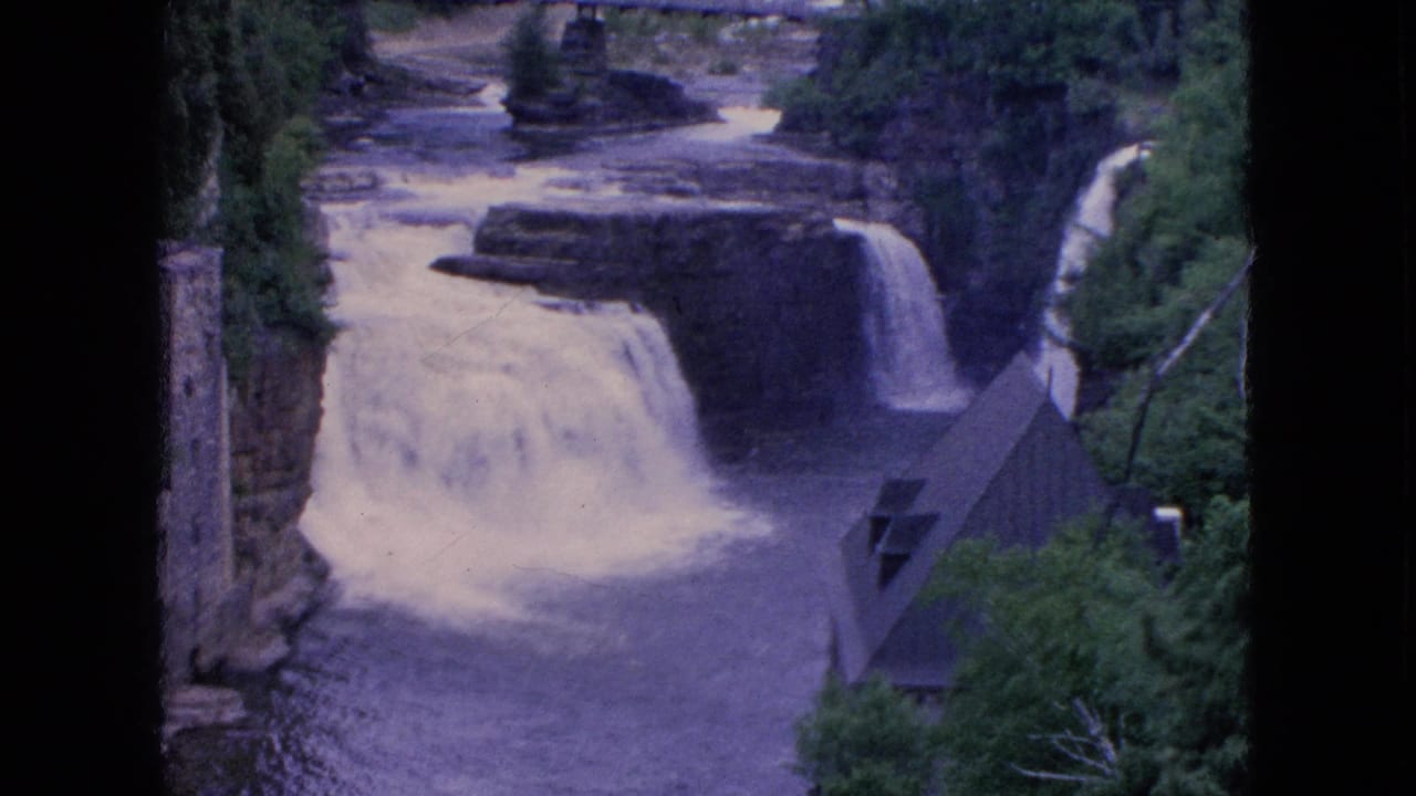 1964 8mm frame from Niagara Falls, New York showing water flowing over rocky ledges into a river, with trees, a stone structure, and a steep-roofed building nearby.