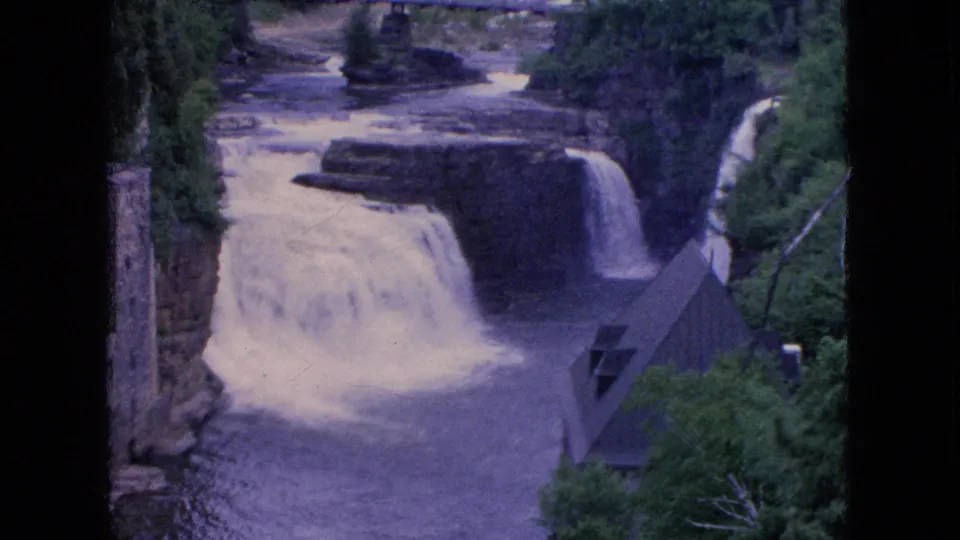 1964 8mm frame from Niagara Falls, New York showing water flowing over rocky ledges into a river, with trees, a stone structure, and a steep-roofed building nearby.