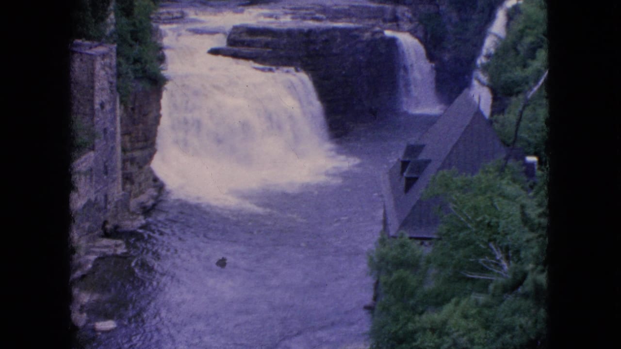 1964 Niagara Falls area home-movie frame showing a waterfall spilling into a dark river, with wooded banks, a stone wall, and a steep-roofed structure at right.