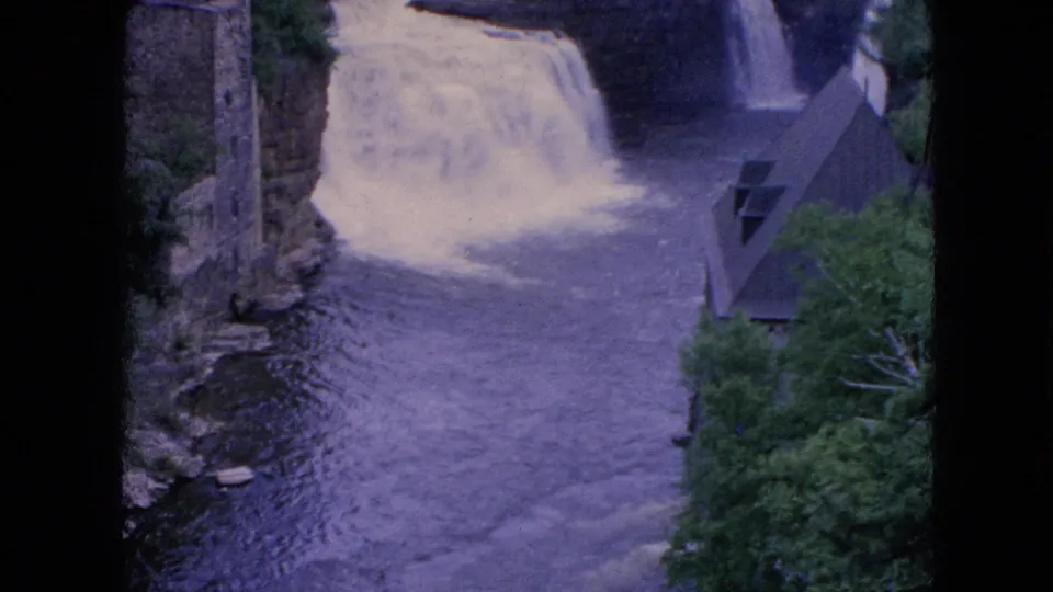 1964 8mm home-movie view of rushing water below a waterfall, framed by rocky banks, trees, a stone structure, and a small angular building.