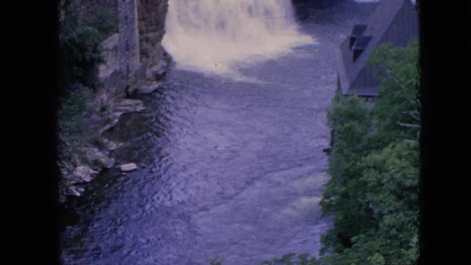 Late frame from the 1964 Niagara Falls clip showing the river below the waterfall, with rippled water, surrounding trees, a stone edge, and a steep-roofed riverside building.