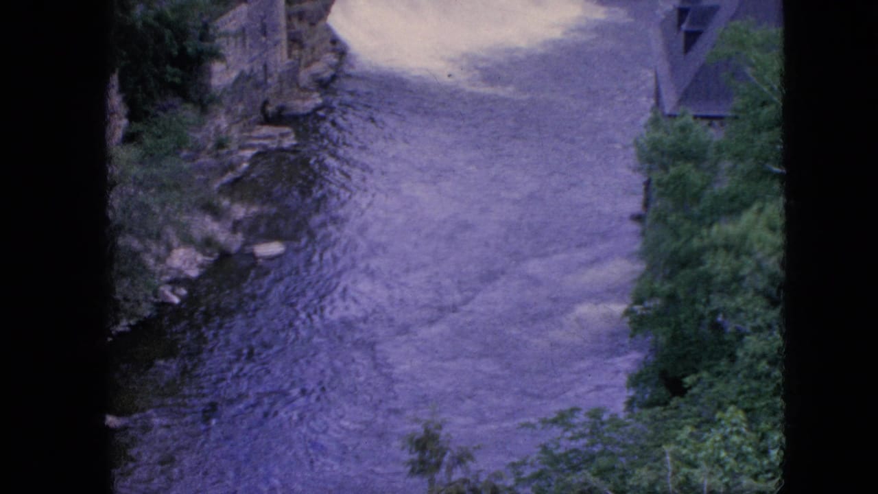 1964 8mm home-movie frame from the Niagara Falls, New York area showing rippled river water flowing between rocky banks with green trees at the right edge.