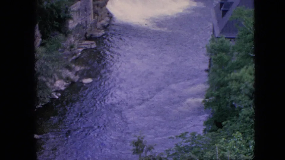 1964 8mm home-movie frame from the Niagara Falls, New York area showing rippled river water flowing between rocky banks with green trees at the right edge.