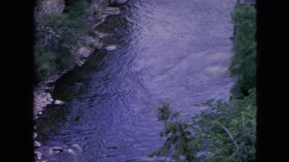 1964 Niagara Falls area home-movie frame showing purple-blue river water moving downstream between a rocky edge and riverside greenery.