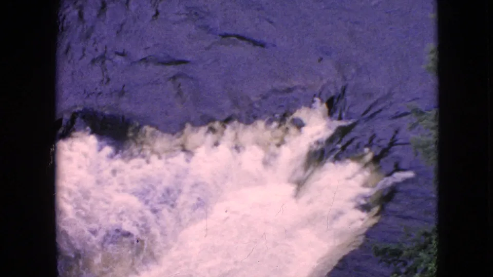 1964 8mm home-movie frame from the Niagara Falls, New York area showing dark river water breaking into frothy white water near a waterfall edge.