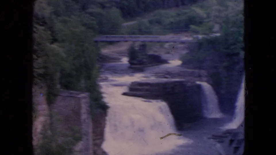 1964 8mm home-movie frame widening to show Niagara Falls water dropping through a rocky channel with a bridge and surrounding greenery in the distance.