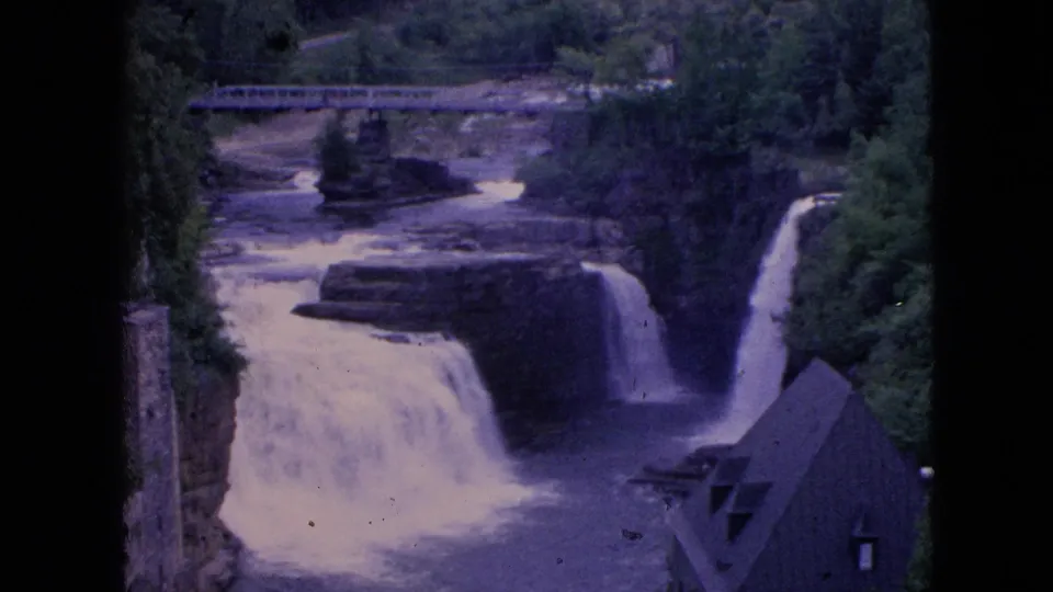 Wider 8mm frame of Niagara Falls showing multiple rapids, a bridge, dark rock ledges, trees, and a roofline near the lower right edge.