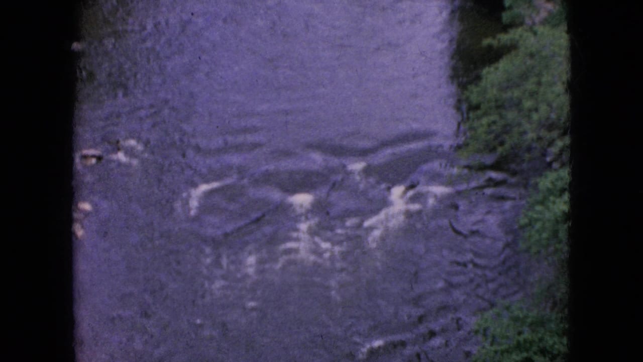 1964 8mm home-movie frame from the Niagara Falls, New York area showing dark rippling stream water beside a small patch of green bank and visible film-edge borders.