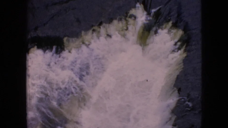 1964 8mm home-movie frame from the Niagara Falls, New York area showing white foaming water rushing through a dark stream, with a small green bank edge and archival film wear visible.