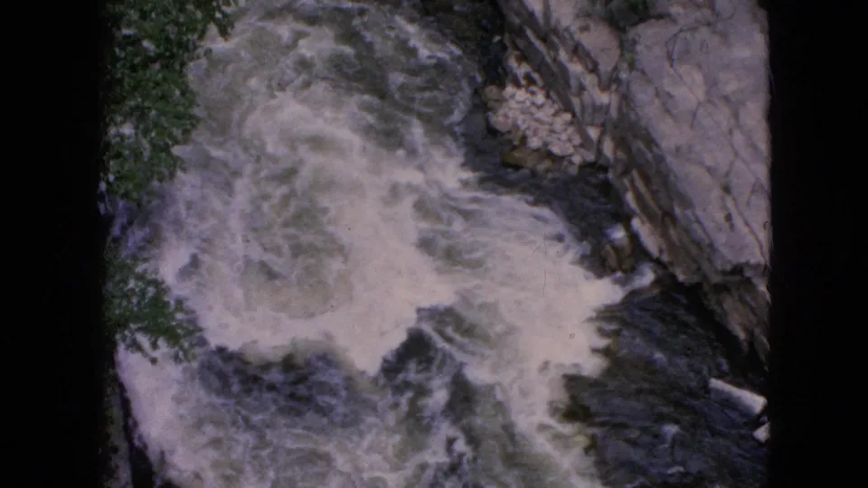 1964 8mm view of Niagara Falls whitewater, with a broad swirl of foam, dark current, green foliage, and a rocky bank along the right side.