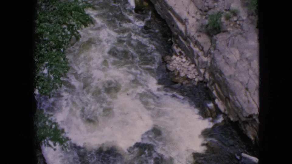 1964 Niagara Falls frame showing frothy rapids moving diagonally through dark water, bordered by rocks and leafy vegetation in a soft 8mm scan.