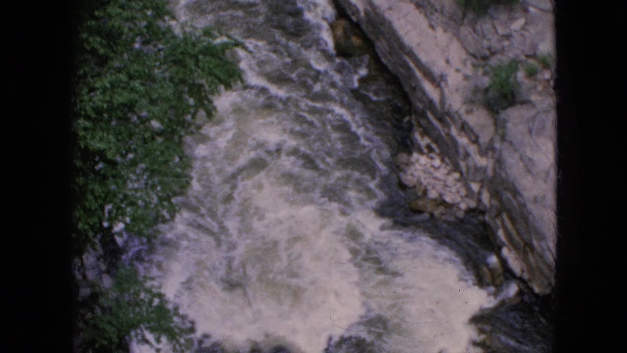1964 8mm home-movie frame of white-water rapids seen from above in the Niagara Falls, New York area, with layered rock and green trees at the canyon edge.