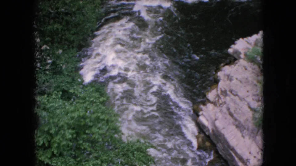 1964 home-movie still showing white-water rapids moving diagonally below trees and exposed rock in the Niagara Falls, New York area.