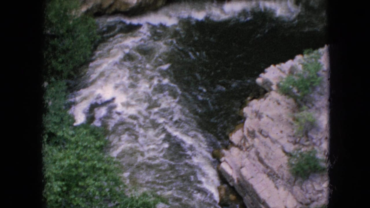 1964 8mm home-movie frame of turbulent water moving through a rocky gorge in the Niagara Falls, New York area, with green foliage along the rim.