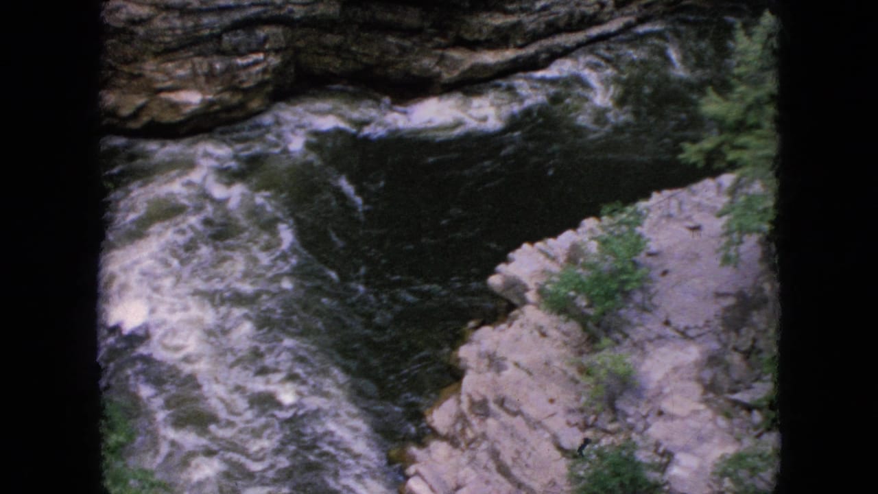 1964 Niagara Falls area home-movie frame showing whitewater swirling beside pale rock ledges and a dark layered cliff face.