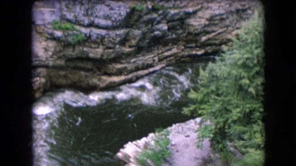 1964 8mm home-movie frame of Niagara Falls area gorge scenery with rushing water, layered rock, green trees, and visible film-frame borders.