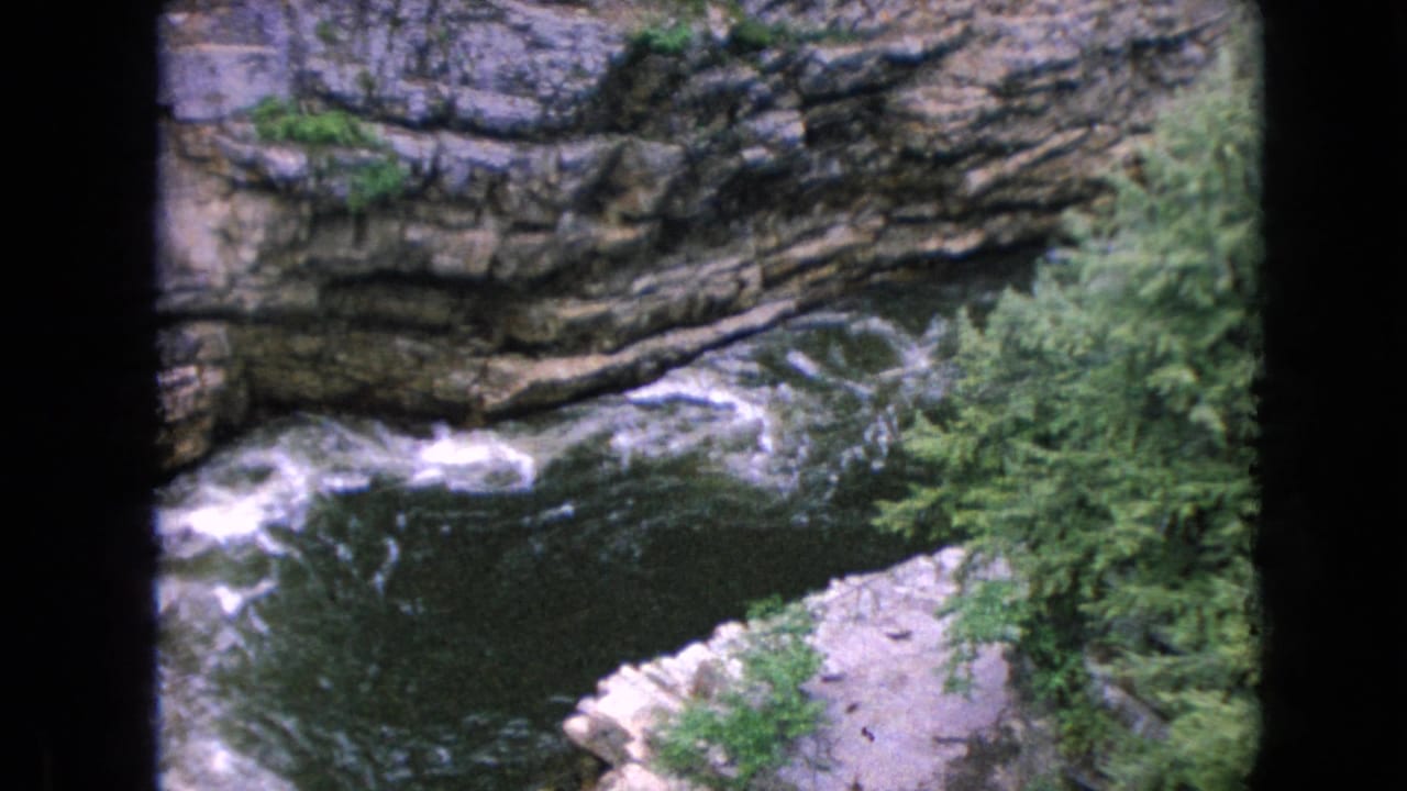 1964 8mm home-movie frame showing a rushing river below layered rock walls and green forest at Ausable Chasm in New York.