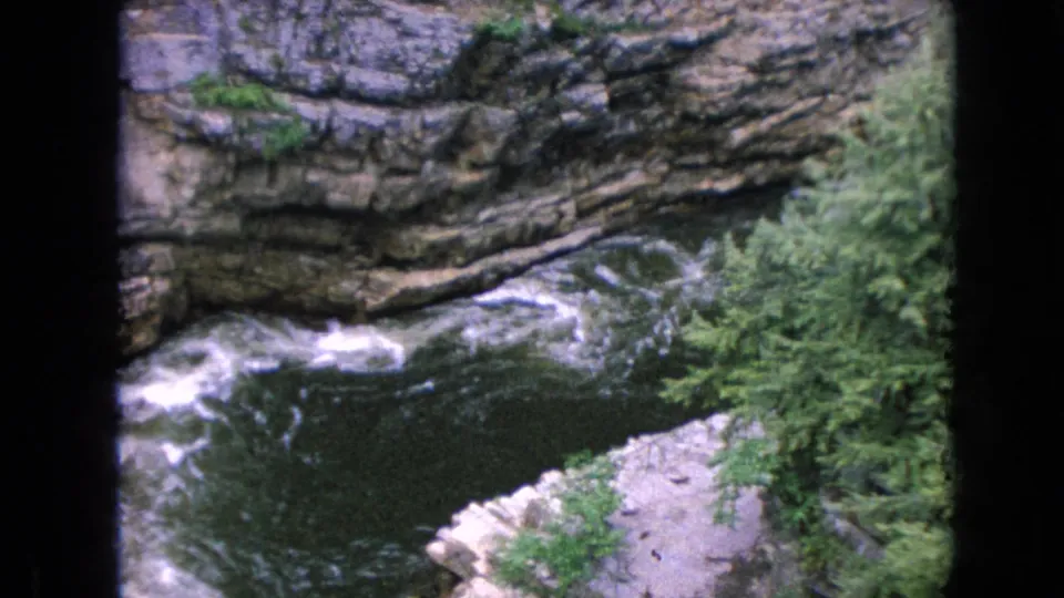 1964 8mm home-movie frame showing a rushing river below layered rock walls and green forest at Ausable Chasm in New York.