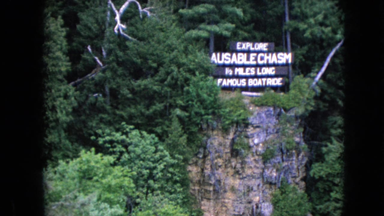 1964 8mm frame with a sign reading Explore Ausable Chasm and 1/2 Miles Long Famous Boatrides set among trees on a cliff face.
