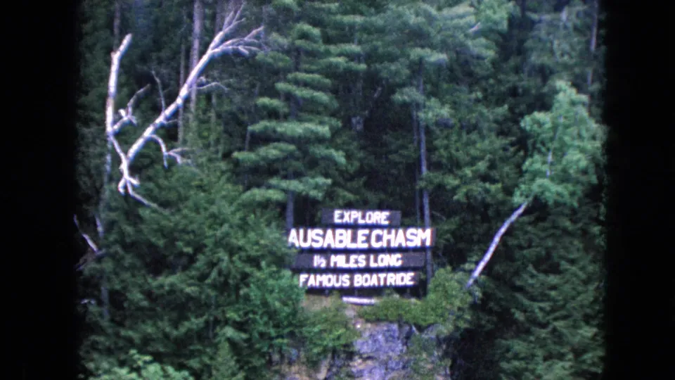 1964 8mm home-movie view of the Ausable Chasm attraction sign surrounded by thick green forest and pale bare branches.
