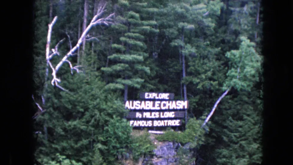 1964 8mm frame showing the Ausable Chasm boat ride sign on a forested cliff, with grainy color and heavy black scan borders visible.