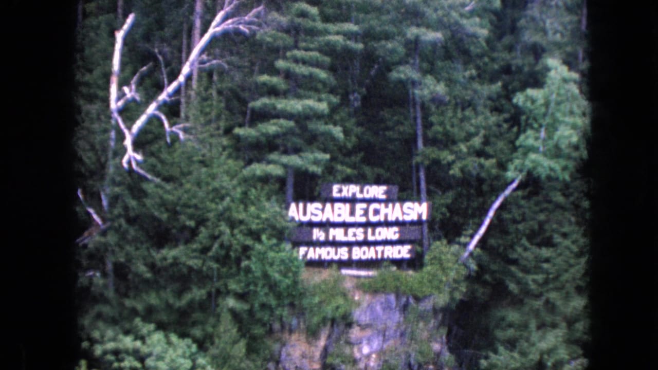 1964 U.S. 8mm frame showing an Ausable Chasm attraction sign set against forest and a rocky cliff, with dark film edges visible.